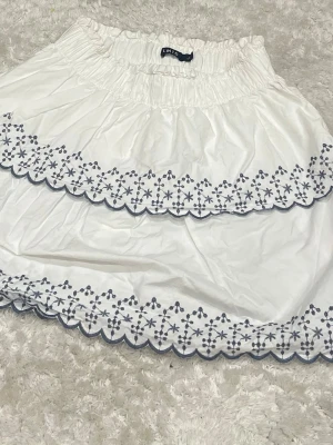 Vit kortkjol med broderad spets - Supersöt vit kortkjol från LMTD med elastisk midja och broderad spetskant i blått. Kjolen har dubbla volanger och ett blommigt broderimönster längs kanterna. Perfekt för dig som gillar en feminin och fräsch stil.