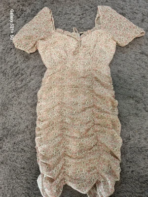 Blommig kortklänning med volang - Söt kortklänning med blommigt mönster i ljusa toner av beige, rosa och grönt. Klänningen har korta puffärmar, volangdetaljer och en liten knytning vid urringningen. Perfekt för sommarens varma dagar.