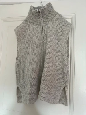 Ljusgrå stickad väst med half zip - Säljer en ljusgrå stickad väst med hög krage och half zip framtill. Västen har ribbad struktur, slitsar i sidorna och är ärmlös. Storleken är L men den passar även M och S ❤️