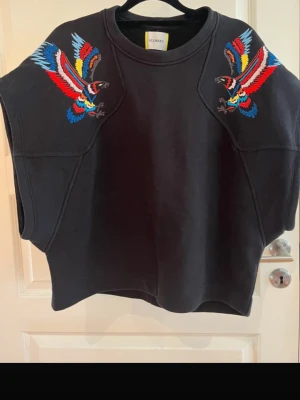 Svart oversized väst med broderi - Cool svart sweatshirt från Iceberg med färgglada broderade fåglar på axlarna i rött, blått och gult. Oversized passform utan ärmar och rund halsringning. Perfekt för dig som vill sticka ut med en unik detalj i din outfit.