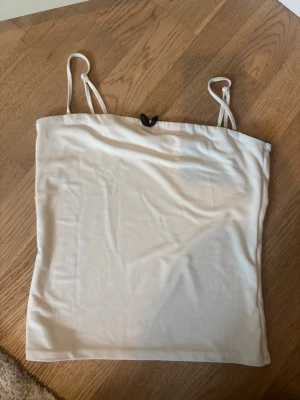 Vit linnetopp med svart rosett - Söt vit linnetopp med tunna axelband och en liten svart rosett framtill. Toppen är enkel och stilren, perfekt att matcha med jeans eller kjol. Materialet känns mjukt och lätt, vilket gör den skön att bära under varma dagar.