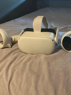 Oculus quest 2 - Säljer ett par vita VR-glasögon med matchande handkontroller. Glasögonen har en mjuk vadderad insida och justerbart band i tyg för bekväm passform. Modern och stilren design, perfekt för gaming och uppslukande upplevelser.