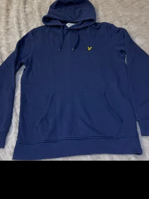 Mörkblå hoodie med gul logga - Snygg mörkblå hoodie med huva och dragsko. På bröstet finns en broderad gul logga. Hoodien har en stor magficka och ribbade muddar vid ärmslut och nederkant. Perfekt för en chill och avslappnad stil.