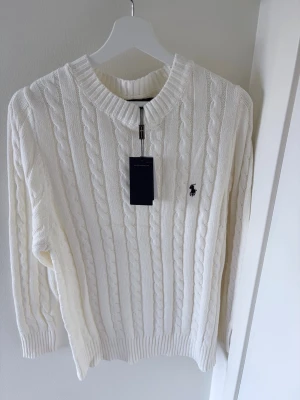 Vit kabelstickad tröja Polo Ralph Lauren - Kabelstickad vit tröja från Polo Ralph Lauren med rund halsringning och broderad marinblå logga på bröstet. Tröjan har ribbade muddar vid ärmslut och nederkant. Klassisk och tidlös design i mjukt material.