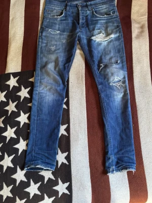 Blå Dondup Jeans  - Säljer ett par blå slim fit jeans med riktigt feta slitningar och hål på benen. Jeansen har klassisk femficksmodell, låg midja och är tillverkade i stretchigt denimtyg för en bekväm passform. Perfekta för en avslappnad och trendig look. Storlek 30. Små defekter finns, fråga om det uppkommer några frågor eller om fler bilder efterfrågas.