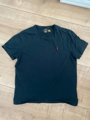 Svart t-shirt med röd logga - En klassisk svart t-shirt i bomull med rund hals och korta ärmar. På bröstet finns en liten röd broderad logga som ger en stilren touch. Perfekt till jeans eller shorts för en enkel och snygg look.