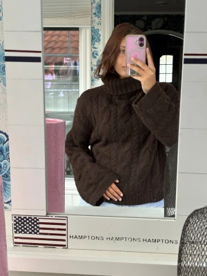 Brun kabelstickad polotröja - Mysig brun kabelstickad polotröja med bred polokrage och långa ärmar. Tröjan har en avslappnad passform och är gjord i ett mjukt, tjockt material som passar perfekt för kyliga dagar. Klassisk kabelstickning ger en tidlös look.