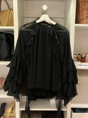 Svart volangblus med vida ärmar - Snygg svart blus i transparent chiffong med volangdetaljer längs ärmar och framstycke. Vida, långa ärmar med flera lager volanger ger en dramatisk look. Perfekt för dig som gillar statement-plagg och flowy material.