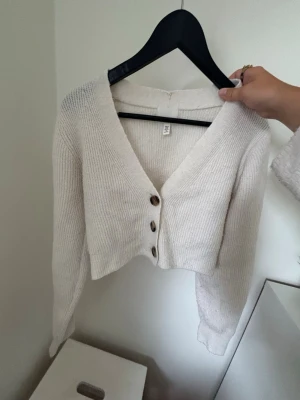Beige cropped kofta med knappar - Säljer en beige cropped kofta från H&M med ribbstickat material och djup V-ringning. Koftan har tre bruna knappar framtill och långa ärmar. Perfekt att slänga över en topp för en chill look.