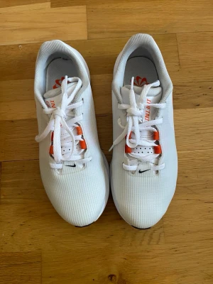 Nike infinity golf sneakers 38.5 - Säljer ett par fräscha vita Nike sneakers med orange swoosh och detaljer. Skorna har snörning och en sportig, modern design med räfflad sula för bra grepp. Ovandelen är i syntetmaterial och mesh för bra andningsförmåga. Perfekta för dig som gillar stilrena och sportiga sneakers.