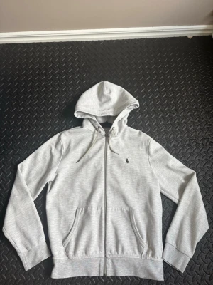 Grå hoodie med dragkedja från Polo Ralph Lauren - Snygg grå hoodie från Polo Ralph Lauren med dragkedja framtill och klassisk logga broderad på bröstet. Hoodien har snörning i huvan, två fickor och ribbade muddar vid ärmar och nederkant. Perfekt för chill dagar eller när du vill vara casual.