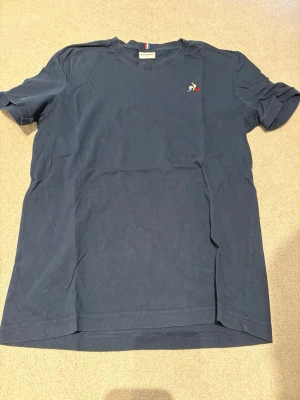 Mörkblå t-shirt med liten logga - En stilren mörkblå t-shirt i bomull med korta ärmar och en liten broderad logga på bröstet. Perfekt till jeans eller shorts för en enkel och clean look. Le coq sportif 