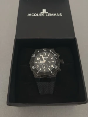 Svart klocka från Jacques Lemans - Snygg svart klocka från Jacques Lemans Champions med sportig design. Klockan har tydliga vita siffror, svart urtavla och robust armband med struktur. Perfekt accessoar för dig som gillar stilrena detaljer och funktionell look kommer med en extra band gratis OBS! Du måste lägga till en batteri eller byta batteri
