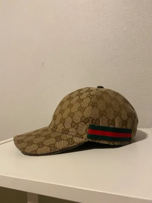 Gucci keps med GG-mönster - Snygg beige keps från Gucci med klassiskt GG-monogram över hela kepsen och en ikonisk grön-röd rand på sidan. Kepsen har böjd skärm och justerbar rem baktill. Materialet är canvas med detaljer i läder.