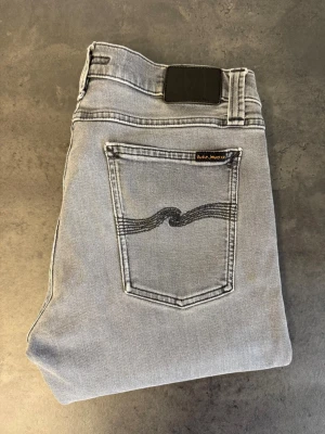 Grå Nudie Jeans, stl 33/32 - Säljer ett par grå jeans från Nudie Jeans med klassisk femficksdesign och svart vågmönster på bakfickan. Jeansen har normal midja och är tillverkade i mjuk denim. Perfekta för en avslappnad och trendig look. NYPRIS:2000kr