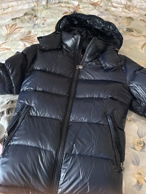 Svart dunjacka från Moncler - Snygg svart dunjacka från Moncler med glansig finish, huva och två dragkedjeförsedda fickor. Jackan har klassisk Moncler-logga på ärmen och breda, vadderade paneler som ger en riktigt puffig look. Perfekt för kalla vinterdagar.