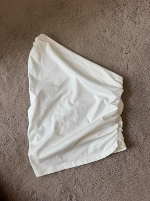 Vit bandeau tubtopp med rynkade sidor - Säljer en stilren vit bandeau tubtopp med rynkade detaljer på sidorna. Toppen är axelbandslös och har en mjuk, stretchig känsla som gör den bekväm att bära. Perfekt att matcha med höga jeans eller kjol för en trendig look.