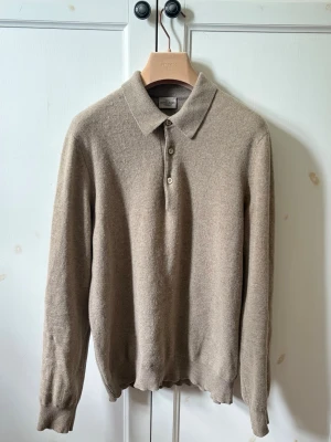 Rose & Born tröja  - Stilren beige långärmad tröja i mjuk cashmere från Rose & Born. Tröjan har klassisk krage och tre knappar framtill, samt ribbade muddar vid ärmslut och nederkant. Perfekt för en avslappnad men ändå uppklädd look.