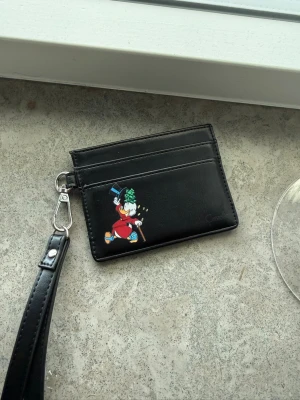 Svart korthållare med lanyard - Snygg svart korthållare i konstläder med motiv av Joakim von Anka som kastar pengar. Har flera kortfack och avtagbar handledsrem i matchande svart. Perfekt för dig som vill ha en lekfull och praktisk accessoar i väskan.