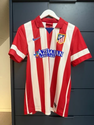 Atlético Madrid matchtröja Villa #9 - Rödvit randig Atlético Madrid matchtröja från Nike med DAVID VILLA och nummer 9 på ryggen. Tröjan har klubbmärke på bröstet, blå Nike-logga och sponsortryck framtill. Tillverkad i lätt och ventilerande material, perfekt för fotboll eller samling.