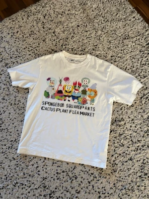 Vit Spongebob x Cactus Plant T-shirt - Vit t-shirt från Uniqlo UT med färgglatt Spongebob Squarepants och Cactus Plant Flea Market-tryck framtill. Klassisk passform med rund hals och korta ärmar. Perfekt för dig som gillar streetwear och unika samarbeten.