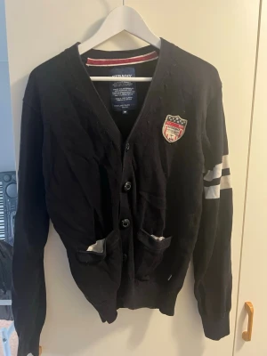 Svart cardigan med emblem - Stilren svart cardigan med knäppning framtill och två fickor. Har vita ränder på ena ärmen och ett broderat emblem på bröstet. Ribbstickade muddar och klassisk passform. Perfekt för en sportig och avslappnad look.