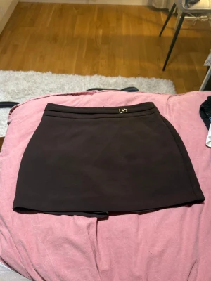 Mörkbrun minikjol - Mörkbrun minikjol med skärpdetalj och guldspänne. Helt oanvänd från NEW YORKER i storlek M. Kort men har insydda shorts. Tajt passform.