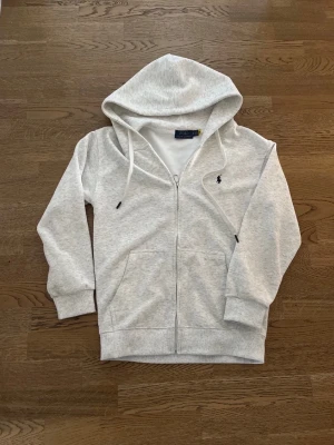 Ljusgrå hoodie med dragkedja - Snygg ljusgrå hoodie från Polo Ralph Lauren med dragkedja framtill och klassisk huva med snören. Har två stora fickor fram och den ikoniska lilla loggan broderad på bröstet. Perfekt för en avslappnad och stilren look. Priset är inte fast!!
