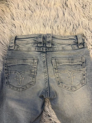 Ljusblå bootcut jeans med slitningar - Säljer ett par ljusblå bootcut jeans med slitningar framtill och snygga broderade bakfickor. Jeansen har klassisk femficksmodell, bälteshällor och är tillverkade i mjuk bomull. Perfekta för en avslappnad och trendig look. Helt nya och oanvända men lappen finns inte kvar då dom är för små