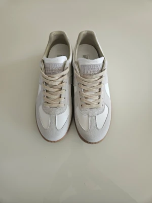 Maison Margiela Gats  - Säljer ett par stilrena sneakers i vitt och beige med ovandel i mocka och släta läderpaneler. Skorna har klassisk låg profil, vita sulor och beige snörning. Perfekta för dig som gillar minimalistisk och clean stil.