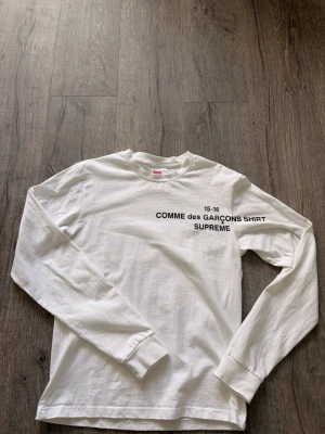 Vit långärmad Supreme x Comme des Garçons - Vit långärmad t-shirt från Supreme och Comme des Garçons med svart tryck på bröstet. Klassisk passform och rund halsringning. Perfekt för dig som gillar streetwear och vill ha något stilrent men ändå unikt.