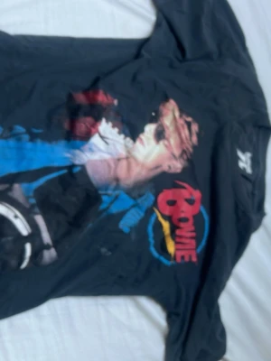 Svart Bowie långärmad t-shirt - Cool svart långärmad t-shirt med stort Bowie-tryck framtill och färgglada detaljer i rött, blått och gult. Baksidan har texten 'North American Tour 74' och turnéstäder i blått och gult. Perfekt för dig som gillar retro och musik.