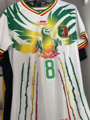 Mali fotbollströja Bissouma - Oanvänd Snygg vit Mali landslagströja från Airness med grönt, gult och rött mönster. Stort örnmotiv på bröstet, nummer 8 och texten 'Bissouma' på ryggen. Ribbade detaljer i landslagets färger vid hals och ärmslut. Perfekt för fotbollsfans!