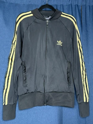Svart Adidas track jacket med guld ränder - Säljer en svart Adidas track jacket med dragkedja och klassiska guld ränder längs ärmarna. Jackan har två fickor framtill, ribbade muddar och Adidas logga på bröstet. Perfekt för en sportig och retro look.🤎 BUDAS UT PÅ LIVE! 🤎 Denna kommer att budas ut på min Plick Live 25 augusti kl. 19:00 🛍️