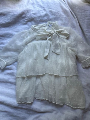 Vit volangklänning med rosett - Söt vit blus med prickigt mönster och volangdetaljer. Blusen har korta ärmar och en stor rosett vid halsen som ger en romantisk vibe. Perfekt för dig som gillar lager-på-lager och en feminin look.