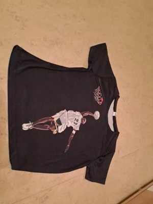 Svart basket t-shirt med tryck - Svart t-shirt i 100% polyester med coolt tryck av en basketspelare som dunkar bollen i korgen. Perfekt för dig som gillar sport och streetstyle. Skön passform och korta ärmar. Säljs för 20 kr. Tveka inte att kontakta om du är intresserad eller har några frågor! Först till kvarn dom gäller!