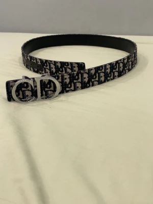 Mönstrat bälte med silverfärgat spänne - Snyggt bälte med svart och grått mönster samt ett iögonfallande silverfärgat spänne. Bältet har ett klassiskt monogramtryck och är tillverkat i textil med insida i svart läder. Perfekt accessoar för att lyfta din outfit.