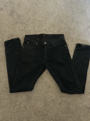 Svarta jeans från Lee W30/L32 - Säljer ett par svarta jeans från Lee med klassisk femficksdesign och knappgylf. Jeansen har smal passform och är tillverkade i stretchigt denimtyg för extra komfort. Perfekta för dig som gillar en stilren och enkel look.