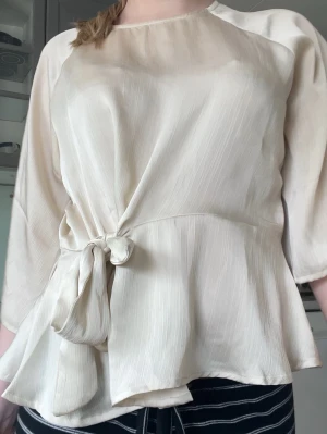 Krämvit blus med knyt i midjan - Snygg krämvit blus i glansigt, lätt material med trekvartsärm och trendigt knyt i midjan. Blusen har en lös passform och subtila vertikala ränder i tyget som ger en elegant känsla. Perfekt att styla med både jeans och kjol. Den har några fläckar i sidan bredvid knytet (sista bilden). 
