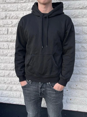 Svart hoodie  - En klassisk svart hoodie med stor magficka och justerbar huva med snören. Hoodien har ribbade muddar vid ärmslut och nederkant. Perfekt för en avslappnad och stilren look. Materialet ser ut att vara mjukt och bekvämt, troligen bomullsblandning. Storlek Xs