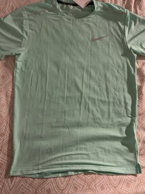 Ljusgrön Nike tränings-tshirt - Snygg ljusgrön tränings-tshirt från Nike med reflekterande detaljer på axlarna och logga på bröstet. Tillverkad i ett lätt och snabbtorkande material som passar perfekt till löpning eller gym. Klassisk rund hals och korta ärmar.