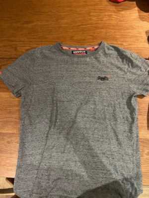Gråmelerad t-shirt från Superdry - Säljer en gråmelerad t-shirt från Superdry med diskret logga broderad på bröstet. Klassisk rund halsringning och korta ärmar. Perfekt basplagg med snygg passform och mjukt material.