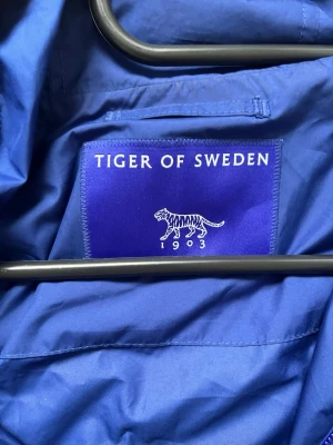 Blå vindjacka från Tiger of Sweden - Snygg blå vindjacka från Tiger of Sweden med huva och dragsko. Jackan har en stor ficka framtill med knappstängning och halv dragkedja. Tillverkad i lätt och tåligt material, perfekt för blåsiga dagar.