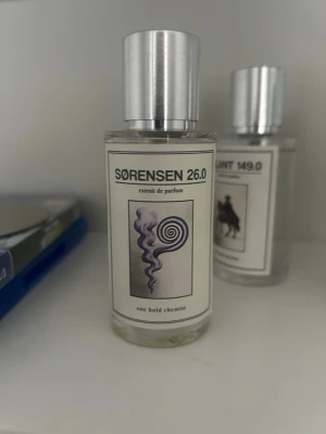 SØRENSEN 26.0 Extrait de Parfum - Unik parfymflaska med minimalistisk design och silverfärgat lock. Flaskan är genomskinlig med en vit etikett och grafiskt motiv i svart och lila. Texten på etiketten är stilren och modern, vilket ger en exklusiv känsla. Perfekt för dig som gillar nischade dofter.