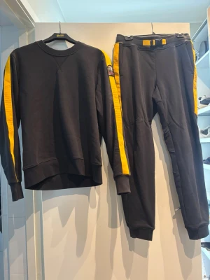 Parajumper set - Snyggt tracksuit set i svart med gula ränder längs ärmar och ben. Setet består av en sweatshirt och matchande byxor. Materialet är mjukt och bekvämt, perfekt för en avslappnad stil. Liten patch på ena ärmen för extra detalj. Ny pris 4500