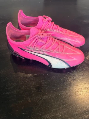 Rosa fotbollsskor från Puma - Snygga fotbollsskor från Puma i en riktigt skarp rosa färg med svarta och vita detaljer. Skorna har låg profil, snörning och mönstrad ovandel i syntetmaterial för bra bollkontroll. Perfekta för dig som vill sticka ut på planen.