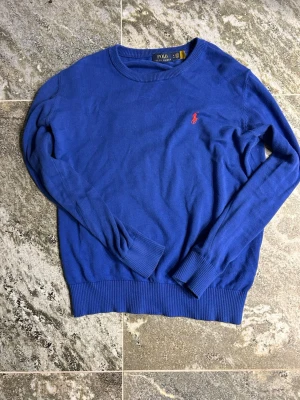 Blå stickad tröja från Polo Ralph Lauren - Klassisk blå stickad tröja från Polo Ralph Lauren med rund halsringning och ribbade muddar. Tröjan har den ikoniska röda logotypen broderad på bröstet. Perfekt till jeans eller kjol för en stilren look.