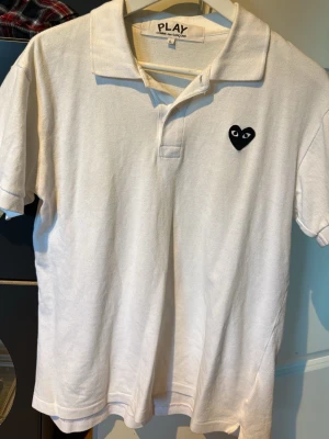 Vit pikétröja Comme des Garçons Play - Vit pikétröja från Comme des Garçons Play med klassisk krage och korta ärmar. Tröjan har den ikoniska svarta hjärtloggan med ögon på bröstet. Perfekt till en avslappnad men ändå stilren look.
