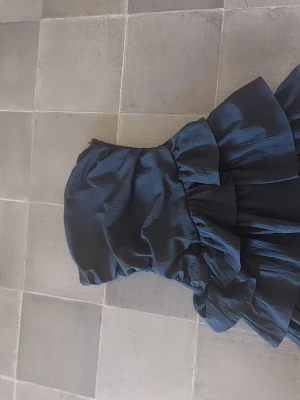 Marinblå axelbandslös volangklänning - Snygg marinblå axelbandslös klänning med volangdetaljer och figurnära passform. Perfekt för dig som vill ha en trendig och elegant look. Klänningen har en kort längd och är tillverkad i ett glansigt material.