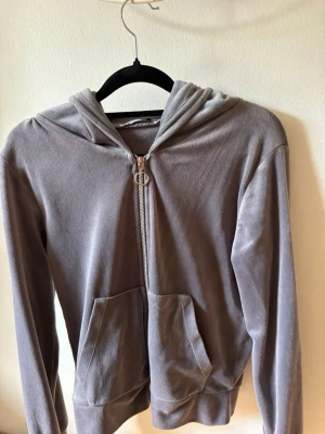 Grå velourhoodie med dragkedja - Säljer en grå hoodie i mjuk velour med dragkedja framtill och två fickor. Hoodien har en klassisk huva och ribbade muddar vid ärmslut och nederkant. Perfekt för en chill och avslappnad stil.